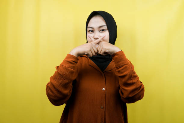Beautiful asian young muslim woman with finger on mouth, telling to be quiet, don't make noise, lower your voice, don't talk, isolated stock photo