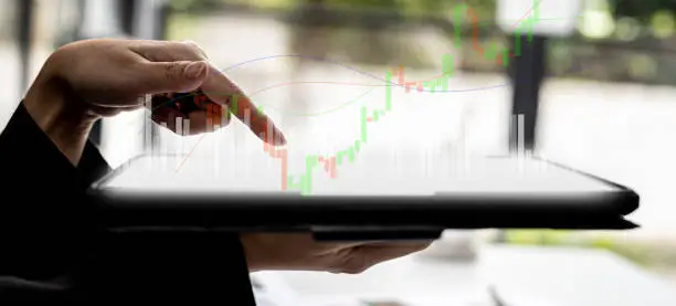 Photo of Person holding a tablet and pointing to a chart diagram showing a stock's up-and-down candlestick chart, graphically displayed on the tablet screen as an uptrend. Stock investor concept.