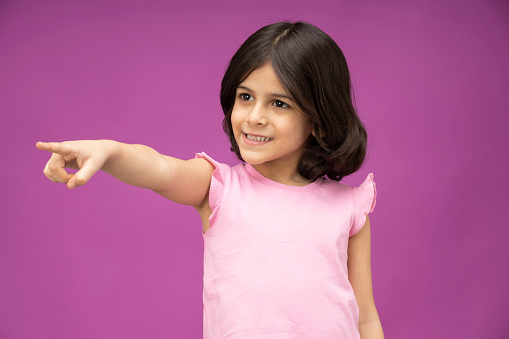 Portrait of cherubic blue-eyed little girl titivating touching short curly fair hair, wearing pink jumpsuit, bracelets, looking at camera, posing on blue background. Childhood, copy space, studio.