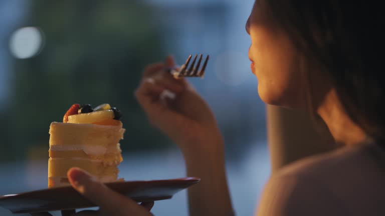 Asian Chinese beautiful woman enjoying slice of cake at home during sunset looking away through window curtain