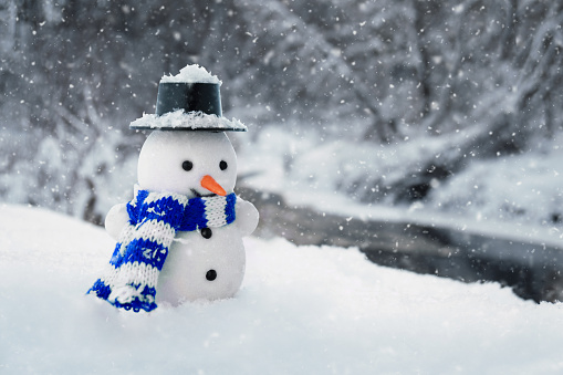 Funny snowman wearing a blue scarf stands in a snowdrift with winter river and forest on the background at countryside in cold snowy day