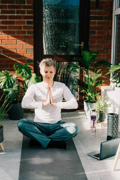 Young man doing yoga position Namaste in cozy loft style room full of home plants on sunny morning. Online class yoga practice. Healthy Lifestyle. Home online workout during a lockdown. Vertical card joven haciendo yoga posición namaste en acogedora habitación estilo loft llena de plantas caseras en la mañana soleada. clase de práctica de yoga en línea. estilo de vida saludable. entrenamiento en línea en casa durante un confinamiento. tarjeta ver