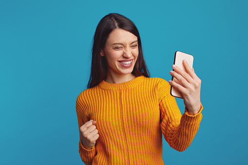 Portrait of delighted woman in yellow sweater clenching fist like winner while using mobile phone isolated over blue background