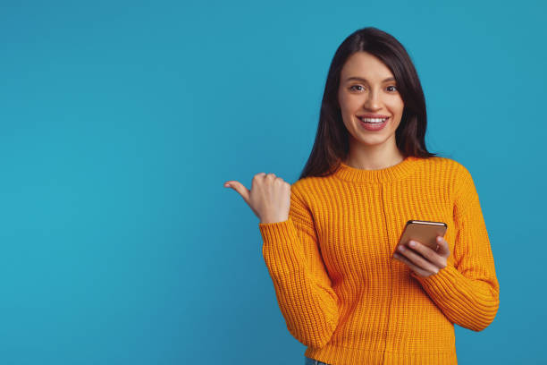 istock Young cute girl pointing at free space while holding smartphone against blue 1359329699