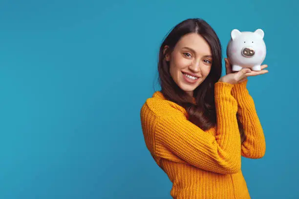 Photo of Young woman in casual orange sweater holding white piggy bank with lots of money