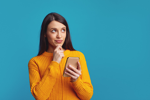 Pretty girl has holds chin thinks about mobile online offer, looks away contemplates about something, isolated over blue background with empty space