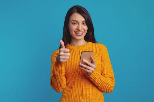 Pretty young girl dressed in warm knitted orange sweater showing thumb up while using smartphone against blue background