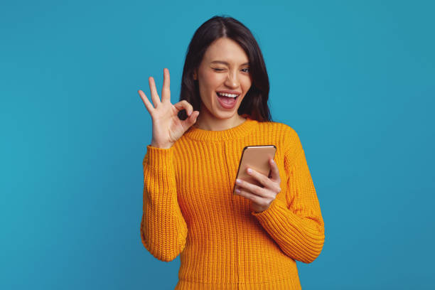 istock Young beautiful woman in orange sweater showing ok gesture while using smartphone 1359330409