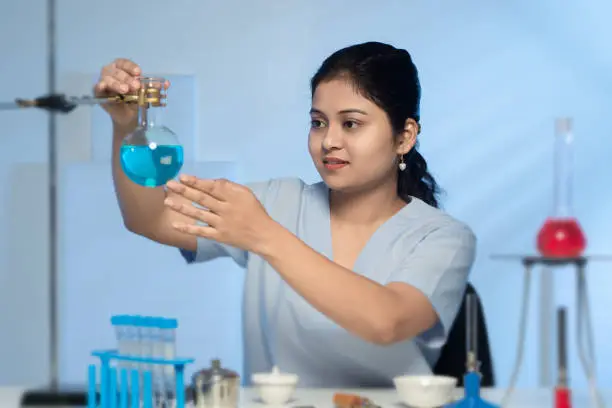 Young Asian Indian Female Lab Technician wearing sky blue uniform, hairs nicely clipped behind, working in laboratory, experimenting, doing research, holding the flask (out of focus) with neck containing blue chemical, thinking and observing closely, with light cyan colour background having rays of shadow from window light giving three-dimensional feel, test tube stand, bunsen burner, spherical flask holder, crucible container, tong, placed on the lab table in the foreground.
