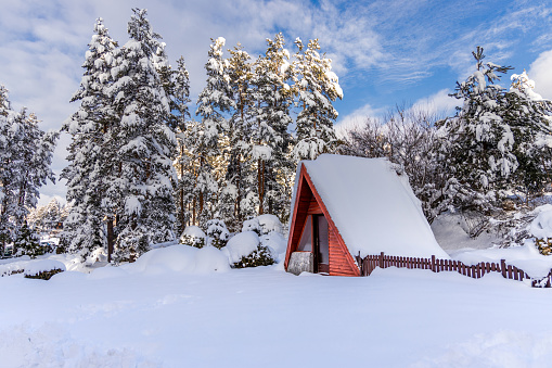 Cute small fairy tale wooden mountain hut covered with snow and surrounded by pine trees. Peaceful isolated winter holiday in A-frame house in scenic nature