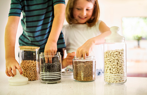 Smiling girl and her little brother taking legumes out of jars sitting on a kitchen counter at home