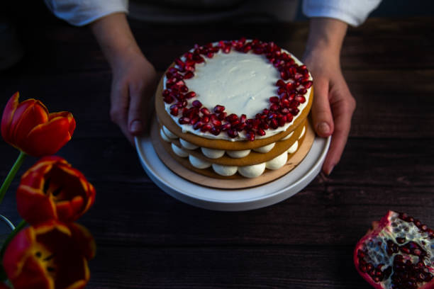 istock The cake stands on a white stand, decorated with cream and pomegranate on top, it is held by the hands of a pastry chef, there is a pomegranate next to it and there are flowers on a dark background 1359344439