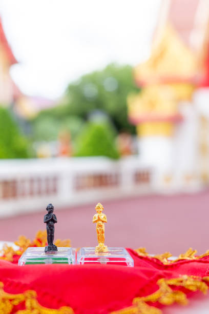 Two of Ai Khai - Kuman Thong, or Thai baby ghost, gold and black amulets standing on white box with red and green velvet pad inside, on red fabric, temple background, focus on foreground, vertical dos de ai khai - kuman thong, o fantasma bebé tailandés, amuletos dorados y negros de pie sobre una caja blanca con almohadilla de terciopelo rojo y verde en el interior, sobre tela roja, fondo del templo, enfoque en primer plano, vertical