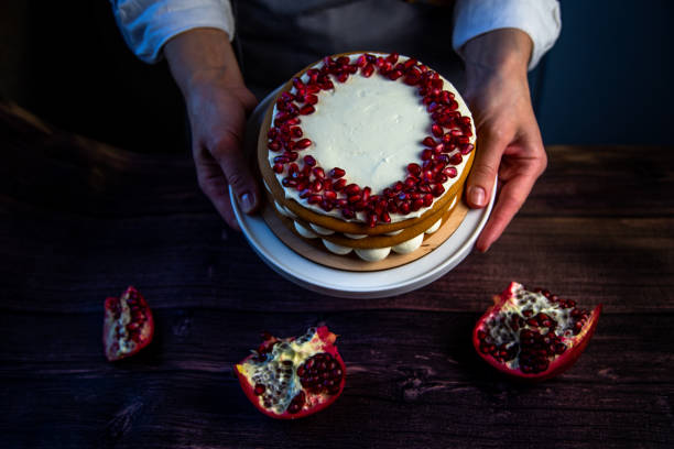 istock A cake with sponge cakes, decorated with white cream and pomegranate seeds on top, is held in a circle by a pastry chef against a background of dark wood and pomegranate 1359348452