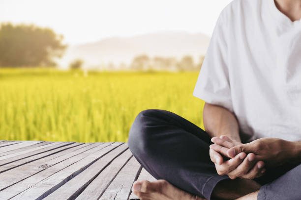 A young man wearing a white T-shirt sitting on old wood bridge is practicing meditation in the morning with the meadow and mountains in the background. stock photo