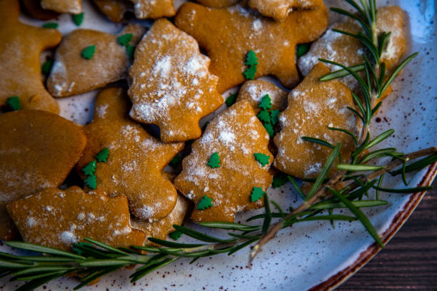 Cookies are on a plate of different shapes, in the foreground there is a cookie in the form of a Christmas tree, decorated with powdered sugar like snow and small green Christmas trees against the background of the rest of the cookies, a branch of rosemary is visible from the side. High quality photo las galletas están en un plato de diferentes formas, en primer plano hay una galleta en forma de árbol de navidad, decorada con azúcar en polvo como la nieve y pequeños árboles de navidad verdes contra el fondo del resto de las galletas, una rama de