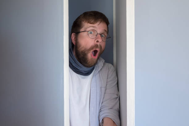 hombre barbudo con ropa casual se esconde en un armario, se asoma por la puerta abierta y mira sorprendido con la boca abierta y los ojos grandes. - hiding fear men peeking fotografías e imágenes de stock