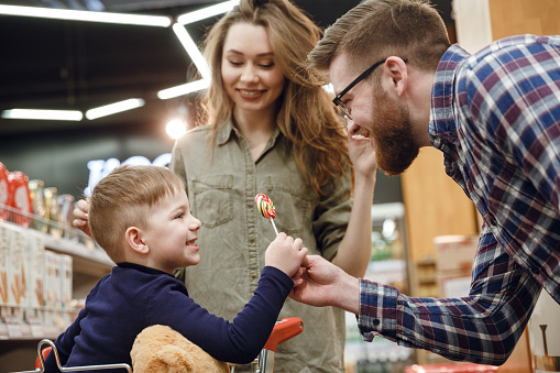 Side view of a bearded father in eyeglasses giving a candy to his son which sitting in shopping trolley in supermarket while mother standing near them
