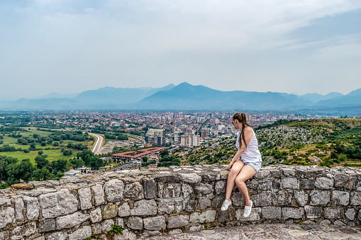 Young adult girl sits on a stone wall of Rozafa Castle and looks at the valley with Shkoder town in the distance, Albania. Woman tourist on the background of a beautiful panorama - view from the back