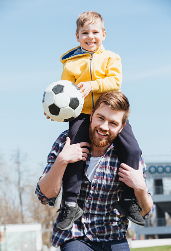 Happy father carrying his little son on shoulders and having fun with a football outdoors