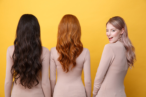 Back view picture of young three ladies standing over yellow background. Blonde cheerful lady winking to camera.