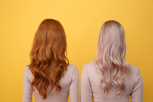 Back view photo of young redhead and blonde ladies friends standing over yellow background.