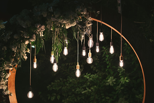 Wedding round arch in rustic style decorated with grass hay field color and retro light bulbs. Near wooden boxes with flower bouquets and candles