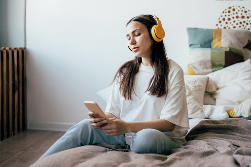 Pregnant attractive young woman sitting on the bed in headphones looks at social networks on a mobile phone.