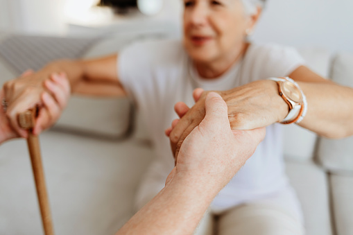 Cropped shot a young male home carer supporting old woman to stand up from the sofa at care home. Photo of male professional caregiver taking care of elderly woman at home during the day.