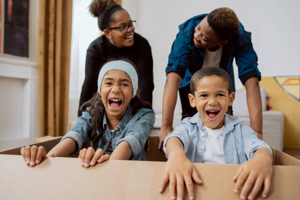 family plays together while moving packing things riding around living room with kids in cardboard box siblings laughing holding each other tight screaming with joy spending time together in apartment - family large american culture fun imagens e fotografias de stock