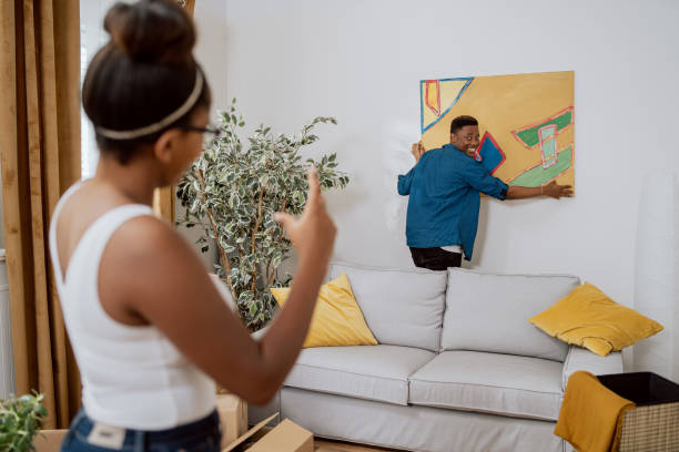 couple amoureux décorant leur premier appartement, déballant les boîtes après avoir emménagé, l’homme accrochant un tableau au mur, la femme montrant combien soulever pour le rendre égal - home decorating photos photos et images de collection