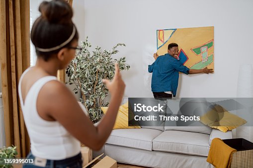 istock Couple in love decorating their first apartment, unpacking boxes after moving in, the man hanging a painting on the wall, the woman showing how much to lift up to make it even 1359395013