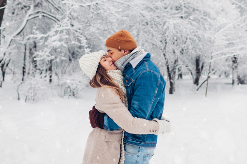 Young stylish loving couple kissing in winter park. Man and woman hugging while walking and enjoying landscape outdoors under falling snow. Valentine's day
