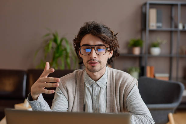 Horizontal portrait of handsome young adult psychologist sitting at desk in his office working with client online on video call using laptop stock photo