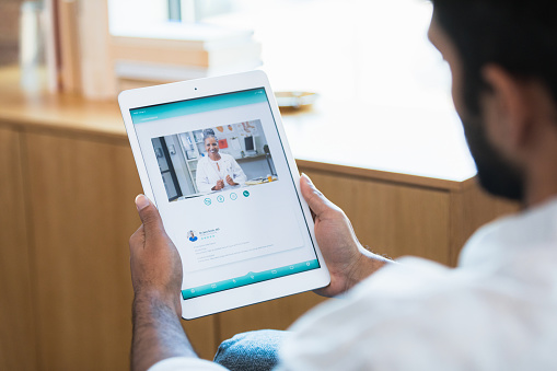 An over the shoulder view of a man sitting in his living room.  He is using a telemedicine app on the digital tablet to consult with his doctor or pharmacist.