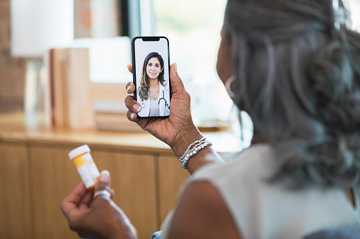 The focus is on the smart phone screen.  The unrecognizable senior adult woman uses a telehealth app to ask her female doctor about the side effects of her new medication.
