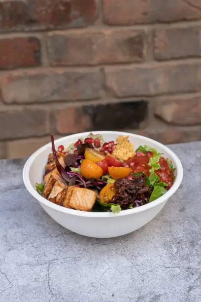 A bowl of grilled chicken salad on a concrete like table and a brick wall in the background.