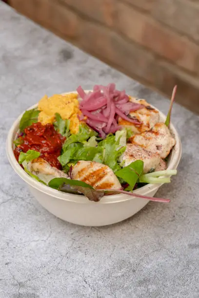 A bowl of grilled chicken salad on a concrete looking table and a brick wall is seen in the background.