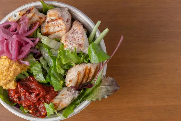 A chicken salad with sauces, onions and green leaves in a takeaway bowl on a wooden table.