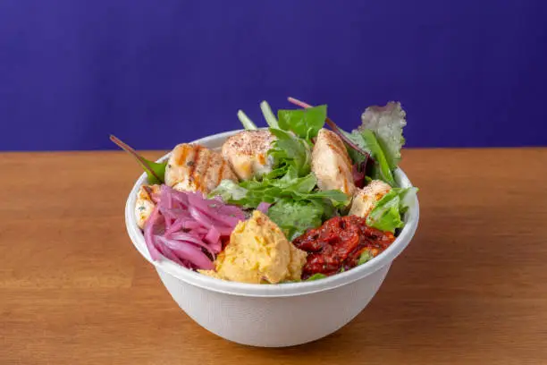 A takeaway bowl of grilled chicken salad with sauces on a wooden table and a purple background.