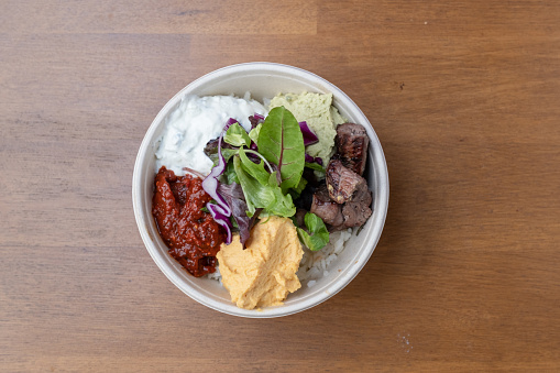 A tabletop view of a takeaway bowl of sish kebab with mezes on a wooden table.