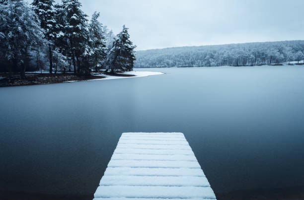 Photo of Dark landscape photo of mole (pier) covered of snow with frozen lake on background - winter time. Frozen and cold lake with hills and forest on background - blue tones.