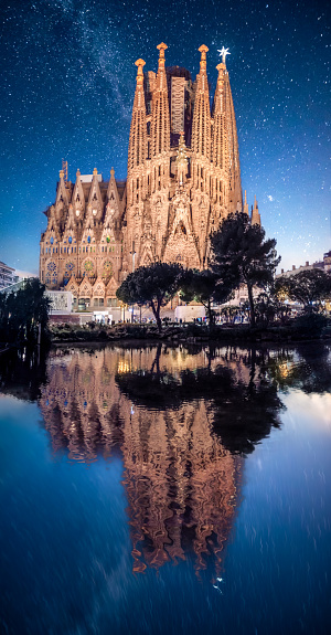 La Sagrada Familia at night reflected in a garden pond in Plaza Gaudi with the Star illuminated Atop The Tower Of The Virgin Mary.