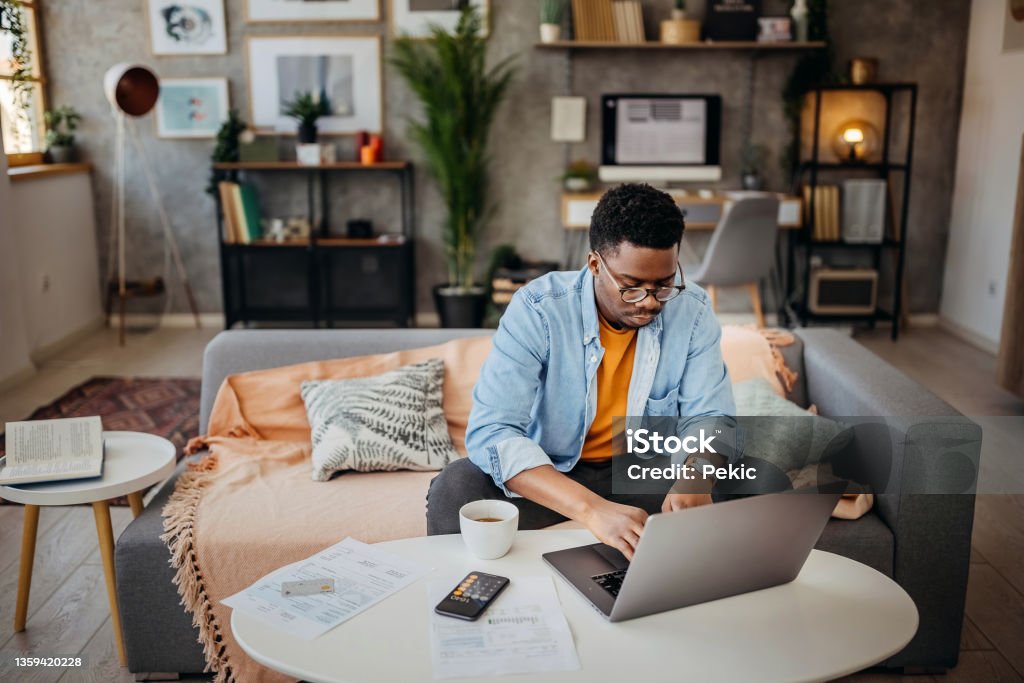 Going over finances at home Young casually clothed man going over finances at his cozy apartment Using Laptop Stock Photo