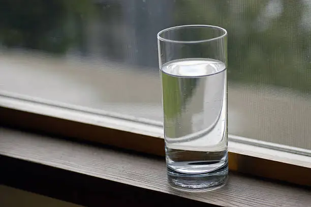 A tall glass of water sits on a window ledge by a window screen on a rainy day