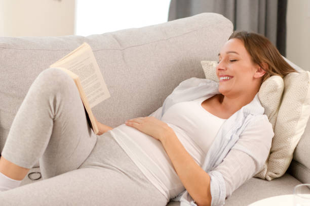 istock Expectant mother reading a book while lying on sofa in living room 1359443286