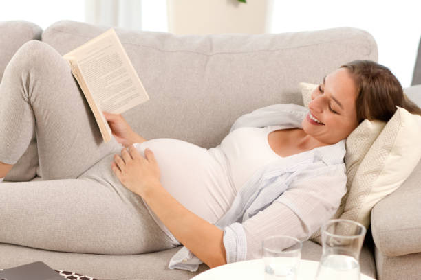 istock Expectant mother reading a book while lying on sofa in living room 1359444378