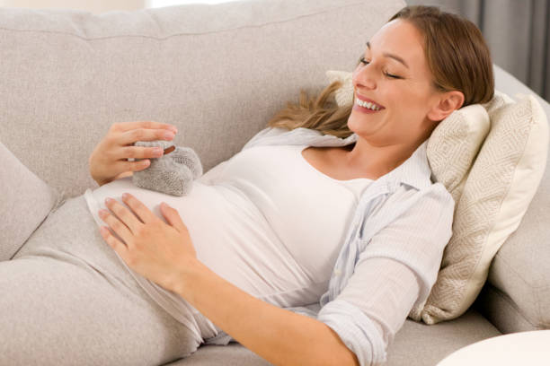 istock Young, pregnant woman showing baby booties while lying down on the sofa 1359444442