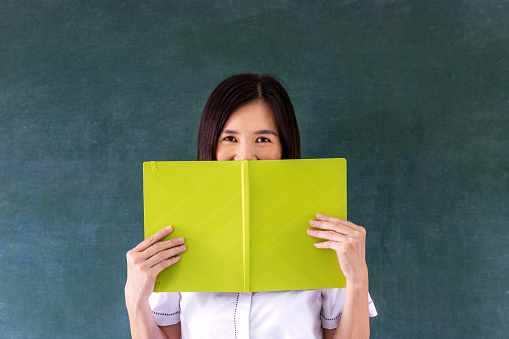 portrait of smart female student bookworm covering face with notebook and stands in front of blackboard