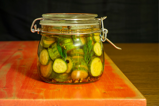 One whole and one halved canned cucumbers on a saucer against the same cucumbers in the sealed glass jar on the rustic table